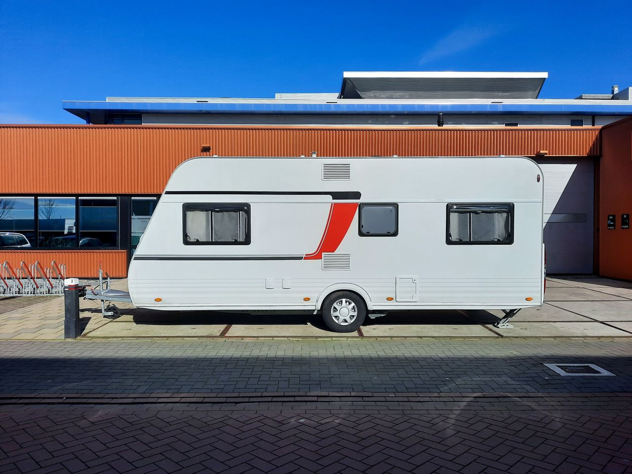 A sleek white caravan with red accents parked next to an industrial building under a clear blue sky.