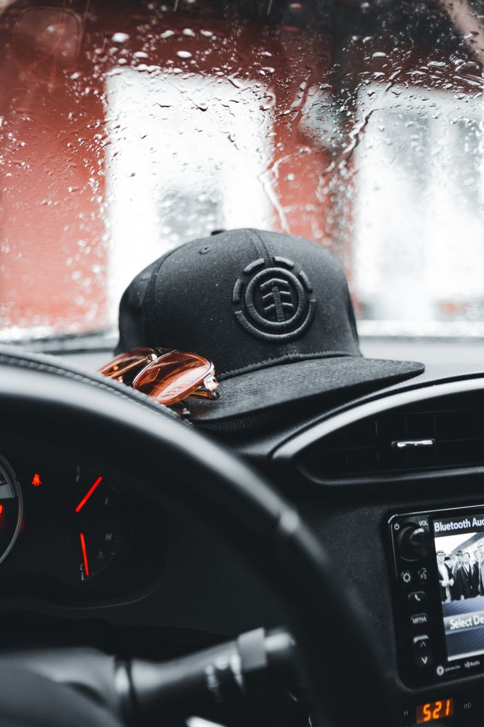 Interior car view with raindrops on the windshield, featuring a black hat and sunglasses on the dashboard.